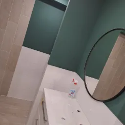 Nowoczesna łazienka po remoncie z białą umywalką nablatową, okrągłym lustrem, zielonymi ścianami i beżowymi płytkami imitującymi drewno, widoczny dozownik mydła i rolka papieru toaletowego.