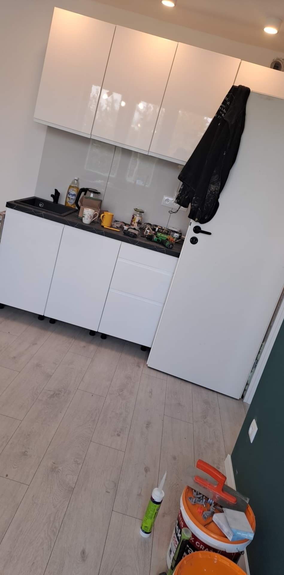 Białe, lakierowane szafki kuchenne z czarnym blatem i zlewozmywakiem, na blacie akcesoria kuchenne i narzędzia, na drzwiach wisi czarna kurtka, na podłodze panele i wiadro z narzędziami.