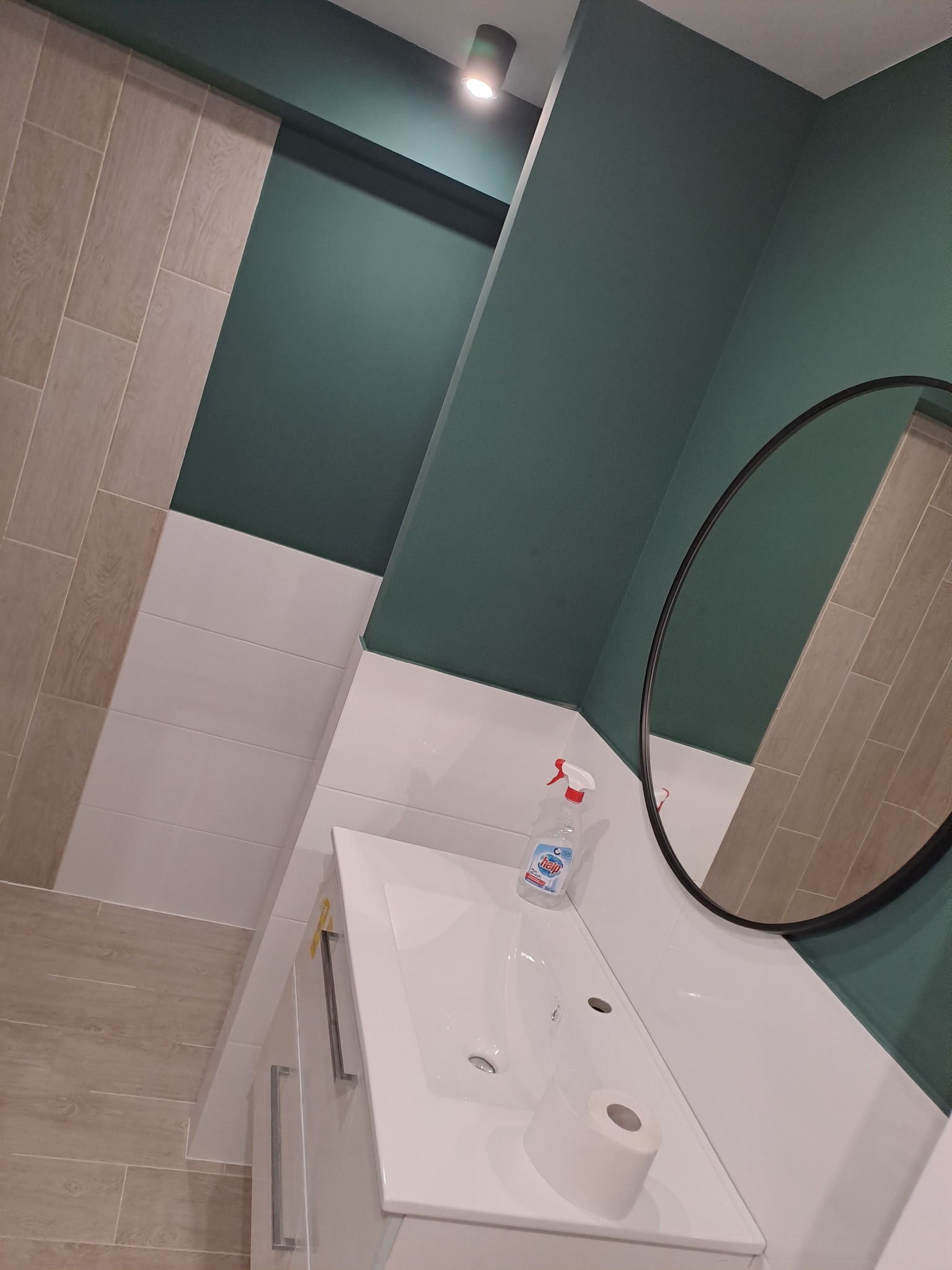 Nowoczesna łazienka po remoncie z białą umywalką nablatową, okrągłym lustrem, zielonymi ścianami i beżowymi płytkami imitującymi drewno, widoczny dozownik mydła i rolka papieru toaletowego.