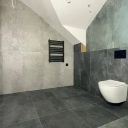 Wykończona łazienka na poddaszu ze ścianami i podłogą wyłożonymi szarymi płytkami imitującymi beton, wisząca toaleta i czarny grzejnik drabinkowy.