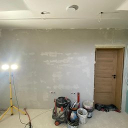 BTC-BUD Firma Remontowo-Budowlana Bartosz Chodura - Pokój w trakcie prac przygotowawczych: ściany zagruntowane, widoczne wyprowadzenia instalacji, sufit podwieszany z otworami na oświetlenie, narzędzia i wiaderka z farbą na podłodze, oświetlenie...