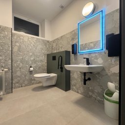 BTC-BUD Firma Remontowo-Budowlana Bartosz Chodura - Nowoczesna łazienka z umywalką, toaletą podwieszaną i lustrem z niebieskim podświetleniem LED. Ściany wykończone płytkami imitującymi lastryko.