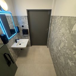 BTC-BUD Firma Remontowo-Budowlana Bartosz Chodura - Nowoczesna łazienka z umywalką, podświetlanym lustrem i płytkami imitującymi lastryko. Minimalistyczny design, szara kolorystyka, widok na drzwi.