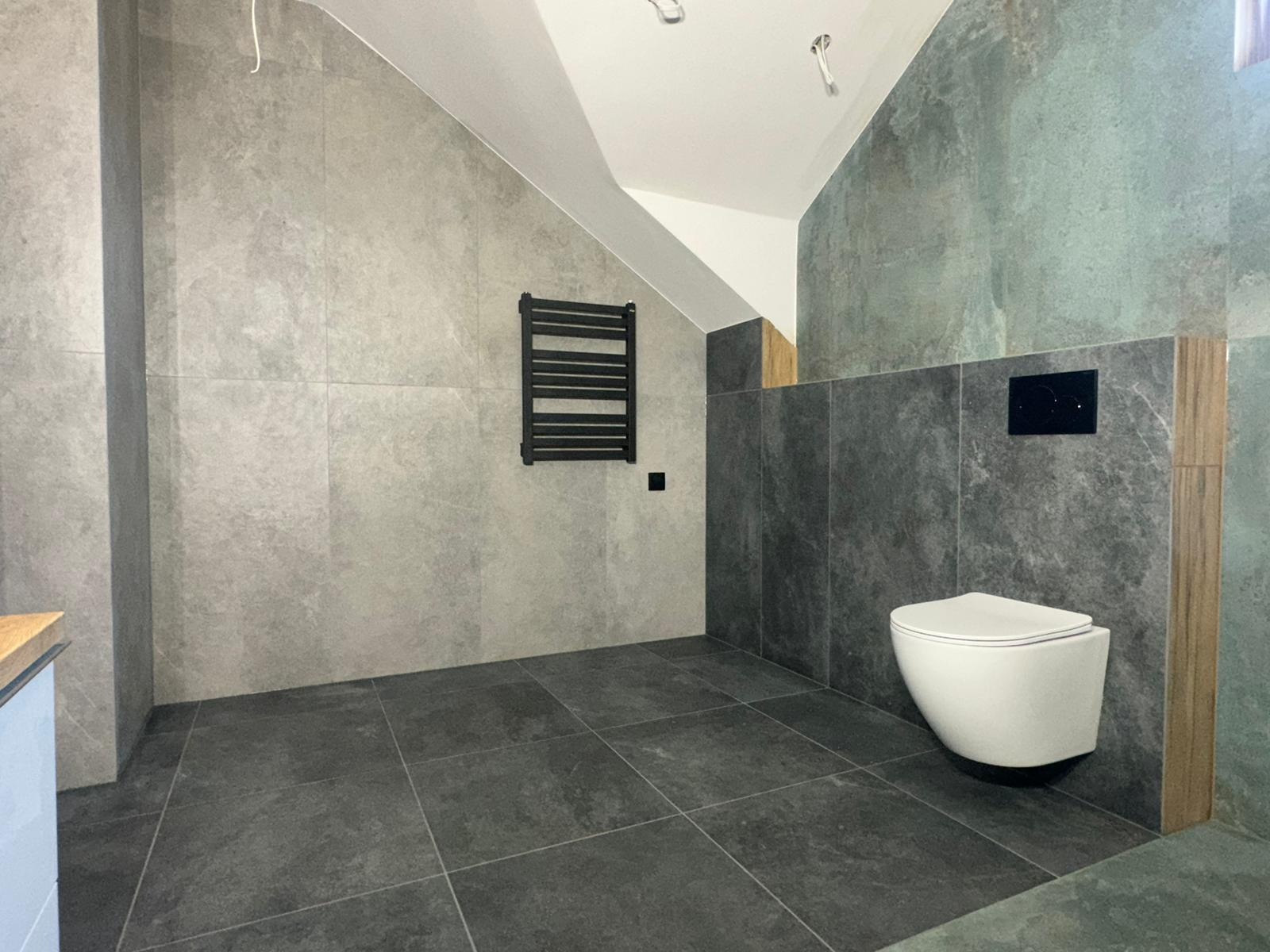 Wykończona łazienka na poddaszu ze ścianami i podłogą wyłożonymi szarymi płytkami imitującymi beton, wisząca toaleta i czarny grzejnik drabinkowy.