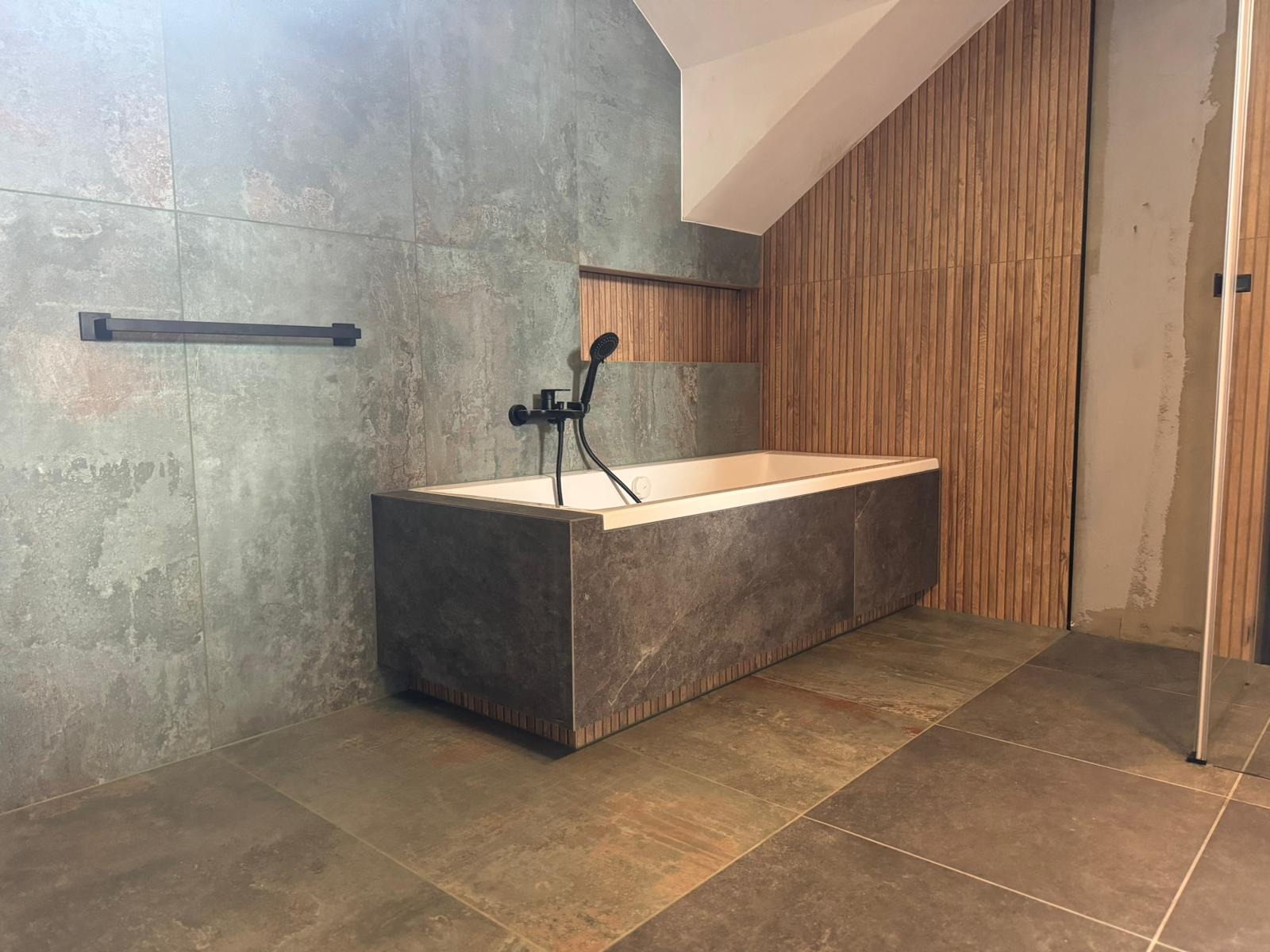 Nowoczesna łazienka z wanną obudowaną płytami imitującymi kamień, czarną armaturą i okładziną ścienną z pionowych drewnianych listew.