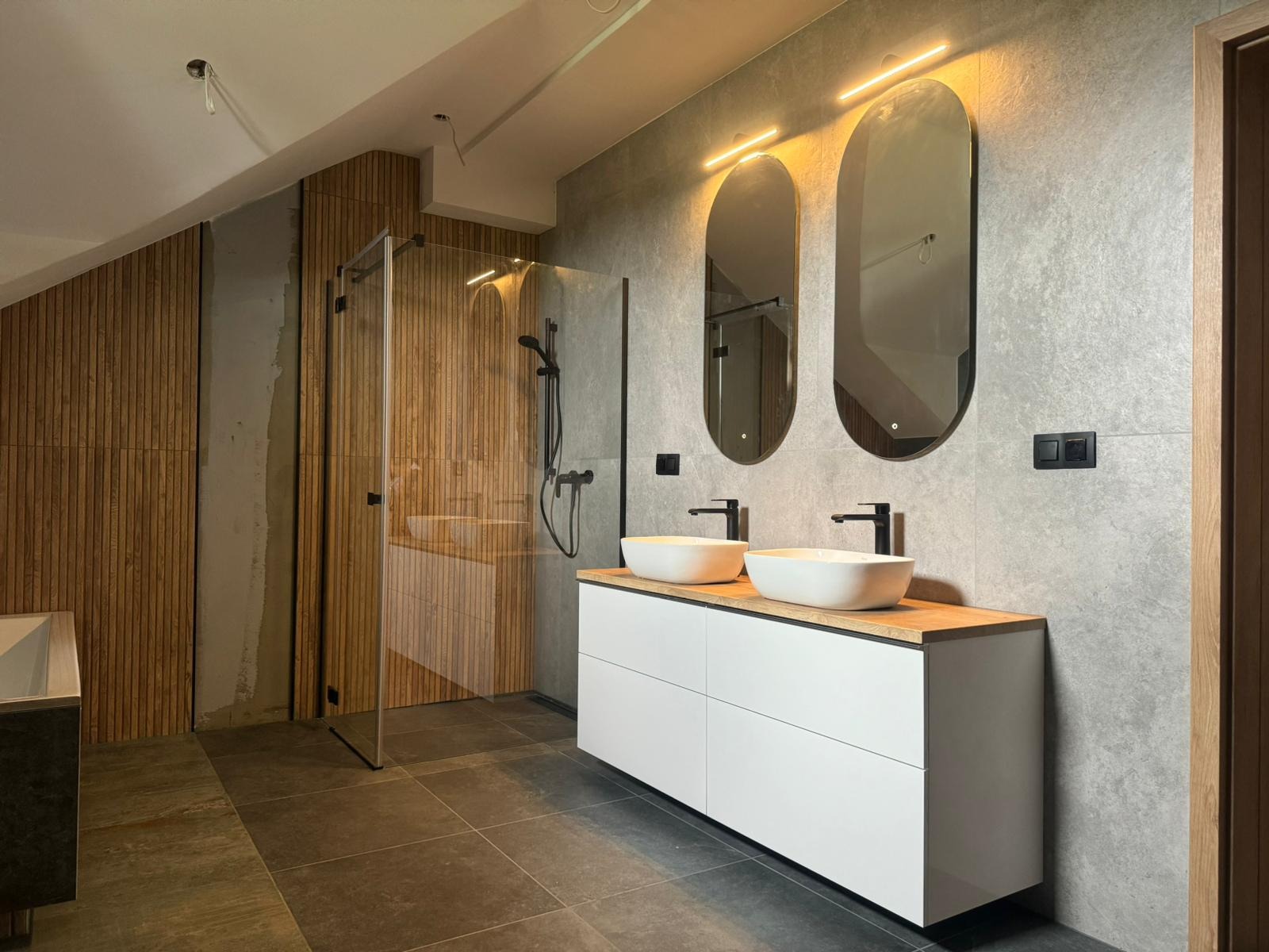 Nowoczesna łazienka na poddaszu z drewnianą ścianą, kabiną prysznicową typu walk-in i podwójną umywalką nablatową na białej szafce z drewnianym blatem, oświetlona lampami nad lustrami.