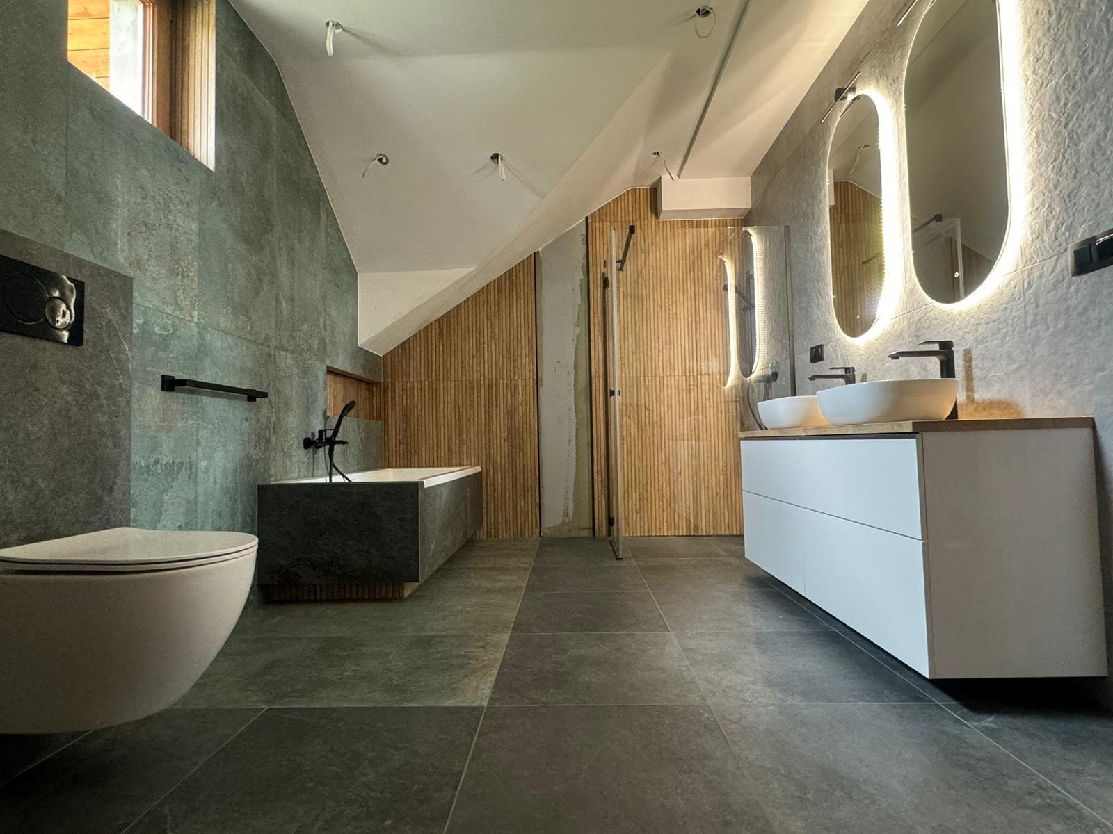 Nowoczesna łazienka na poddaszu z szarymi płytkami, wanną obudowaną kamieniem, białymi szafkami z umywalkami i podświetlanymi lustrami owalnymi.