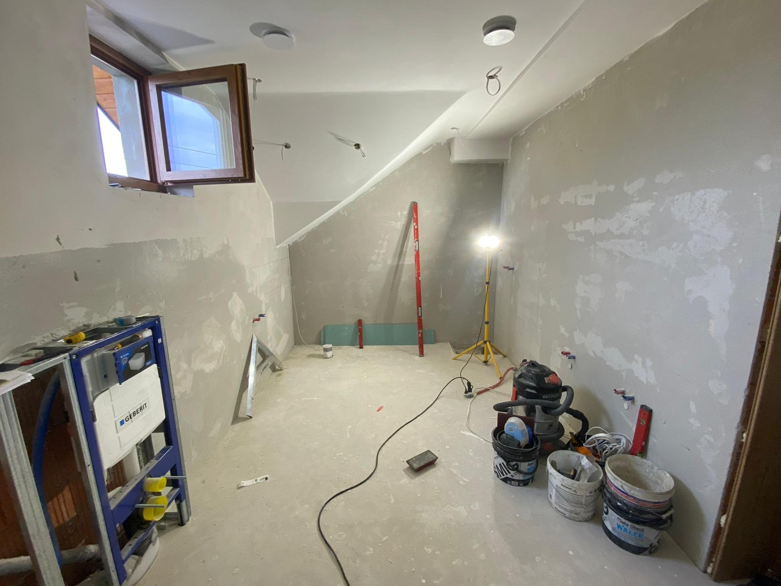 Łazienka w trakcie prac wykończeniowych: widoczny stelaż podtynkowy Geberit, ściany po gruntowaniu, poziomica, płyta gipsowo-kartonowa, lampa budowlana na statywie, odkurzacz przemysłowy i wiadra...