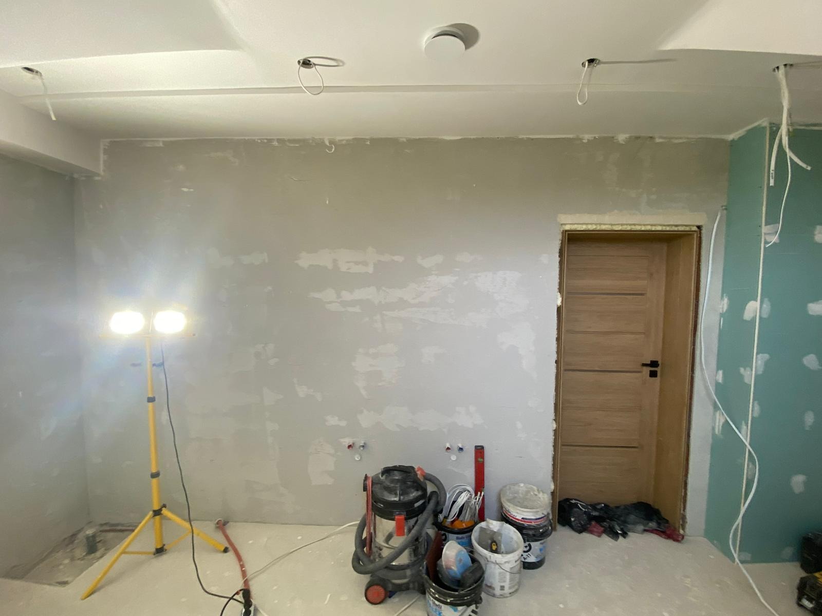 Pokój w trakcie prac przygotowawczych: ściany zagruntowane, widoczne wyprowadzenia instalacji, sufit podwieszany z otworami na oświetlenie, narzędzia i wiaderka z farbą na podłodze, oświetlenie...