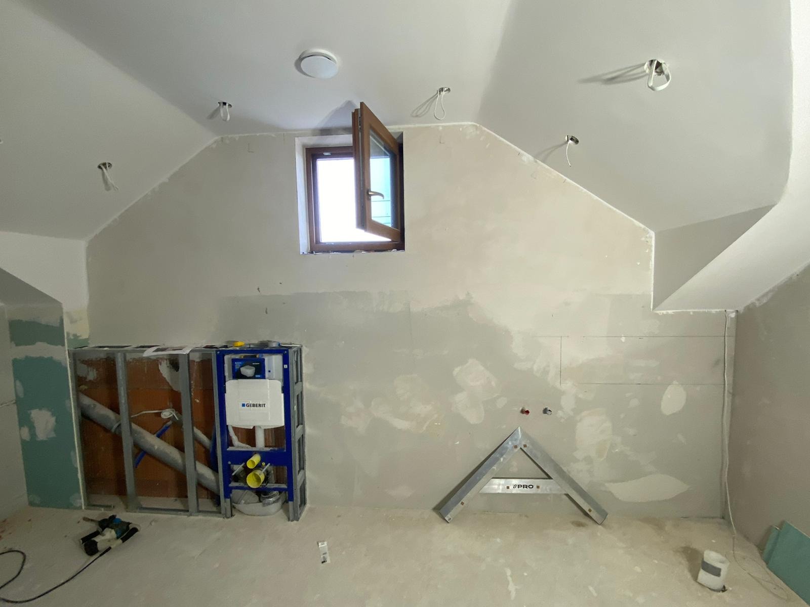 Niedokończona łazienka na poddaszu ze stelażem podtynkowym Geberit, otwartym drewnianym oknem i częściowo położoną płytą karton-gips na ścianach.