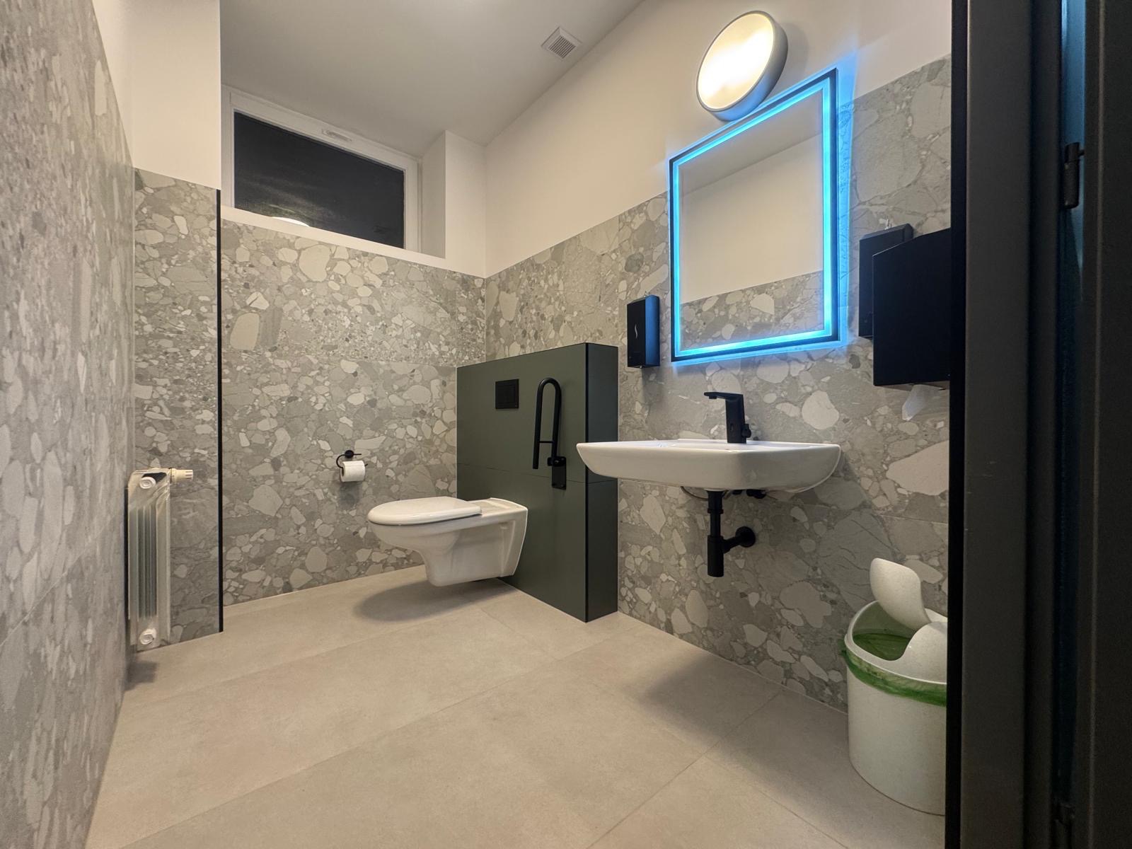 Nowoczesna łazienka z umywalką, toaletą podwieszaną i lustrem z niebieskim podświetleniem LED. Ściany wykończone płytkami imitującymi lastryko.