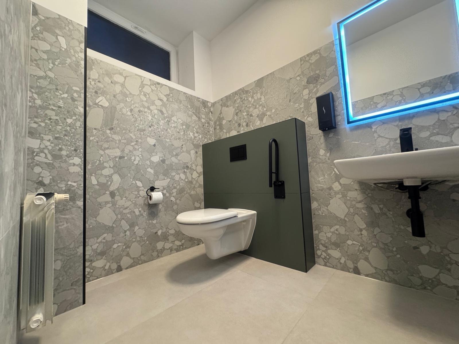 Nowoczesna łazienka z szarą mozaiką na ścianach, podwieszaną toaletą na tle oliwkowozielonej zabudowy i lustrem z niebieskim podświetleniem LED.