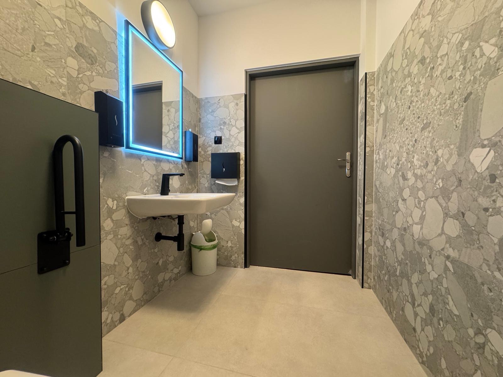 Nowoczesna łazienka z umywalką, podświetlanym lustrem i szarą mozaiką na ścianach. Widoczny kosz na śmieci i uchwyt dla niepełnosprawnych. Minimalistyczny design.