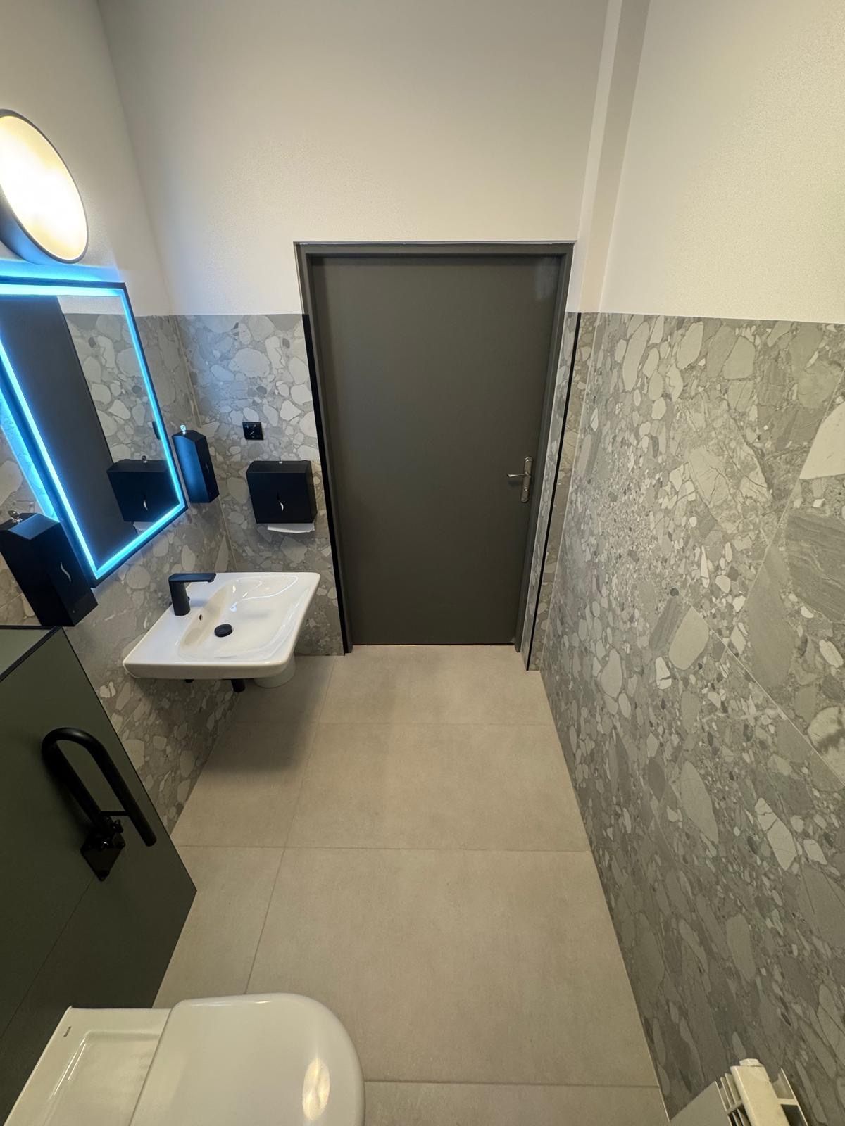 Nowoczesna łazienka z umywalką, podświetlanym lustrem i płytkami imitującymi lastryko. Minimalistyczny design, szara kolorystyka, widok na drzwi.