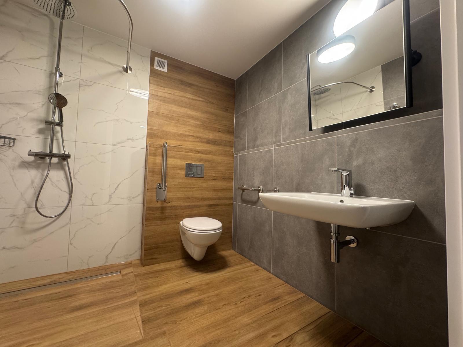 Nowoczesna łazienka z kabiną prysznicową typu walk-in, umywalką nablatową i toaletą podwieszaną. Wykończenie w odcieniach szarości, bieli i drewna. Minimalistyczny design.