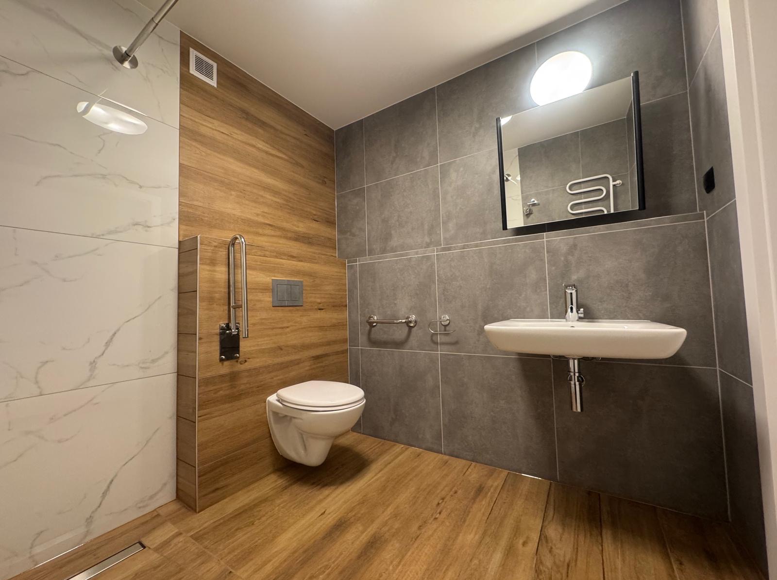 Nowoczesna łazienka z drewnopodobnymi i marmurowymi płytkami, wiszącą toaletą i umywalką. Udogodnienia dla osób z niepełnosprawnościami, minimalistyczny design.