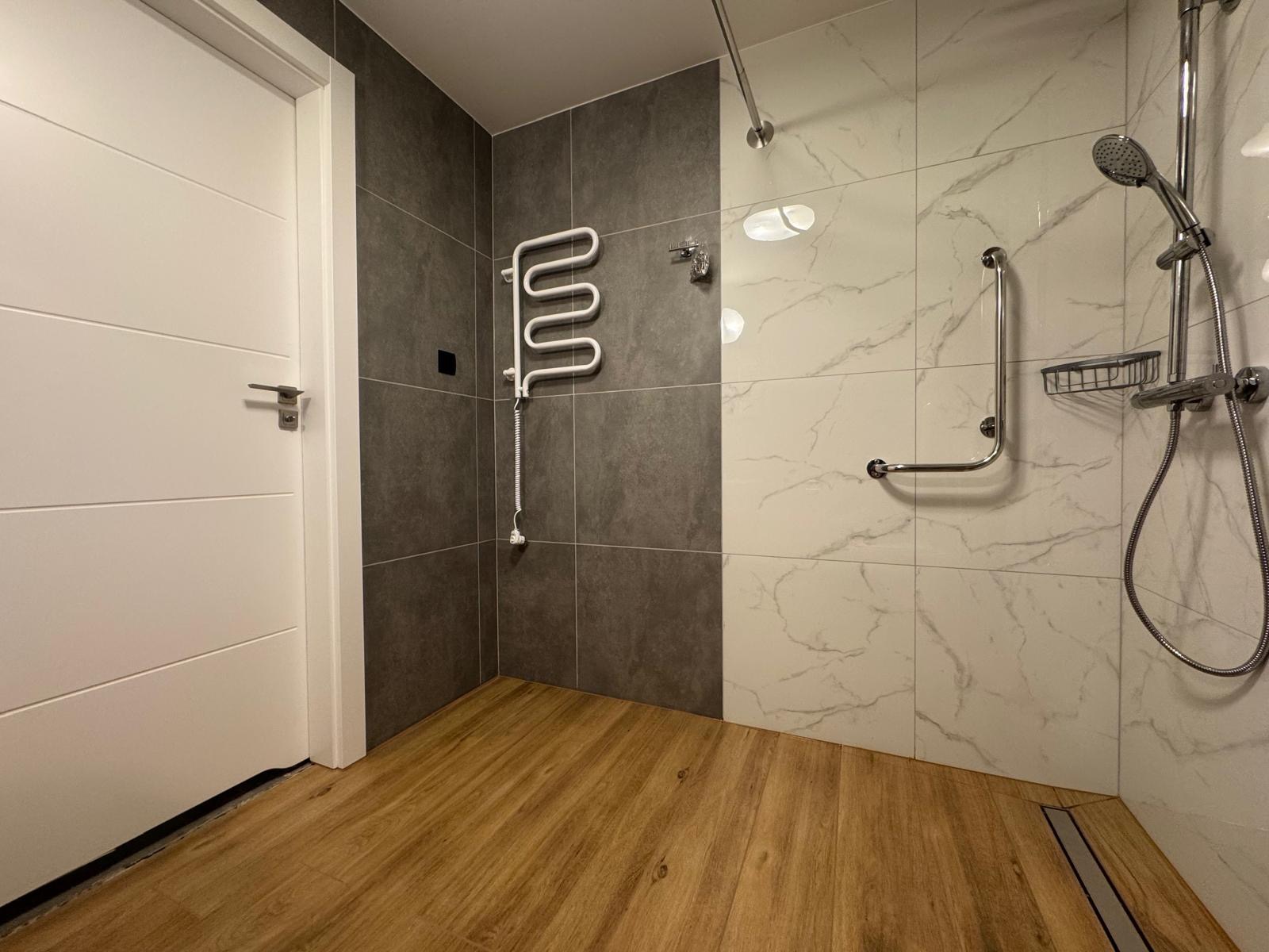 Nowoczesna łazienka z drewnopodobną podłogą, szarymi i marmurkowymi płytkami, grzejnikiem drabinkowym i uchwytem dla niepełnosprawnych przy prysznicu.