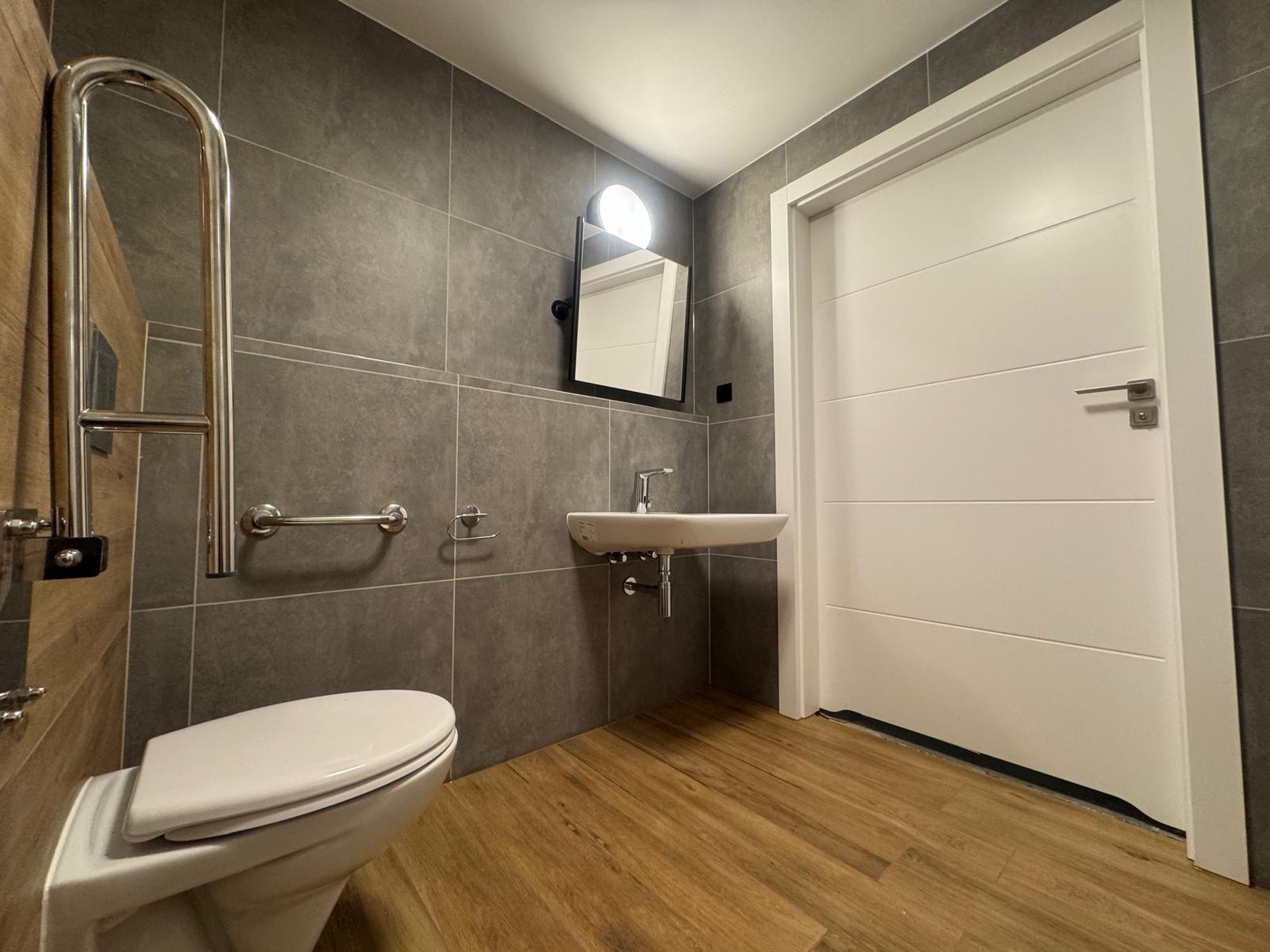 Nowoczesna łazienka z szarymi płytkami, drewnopodobną podłogą i białymi elementami: toaleta, umywalka, drzwi. Widoczne uchwyty dla niepełnosprawnych i lustro z oświetleniem.