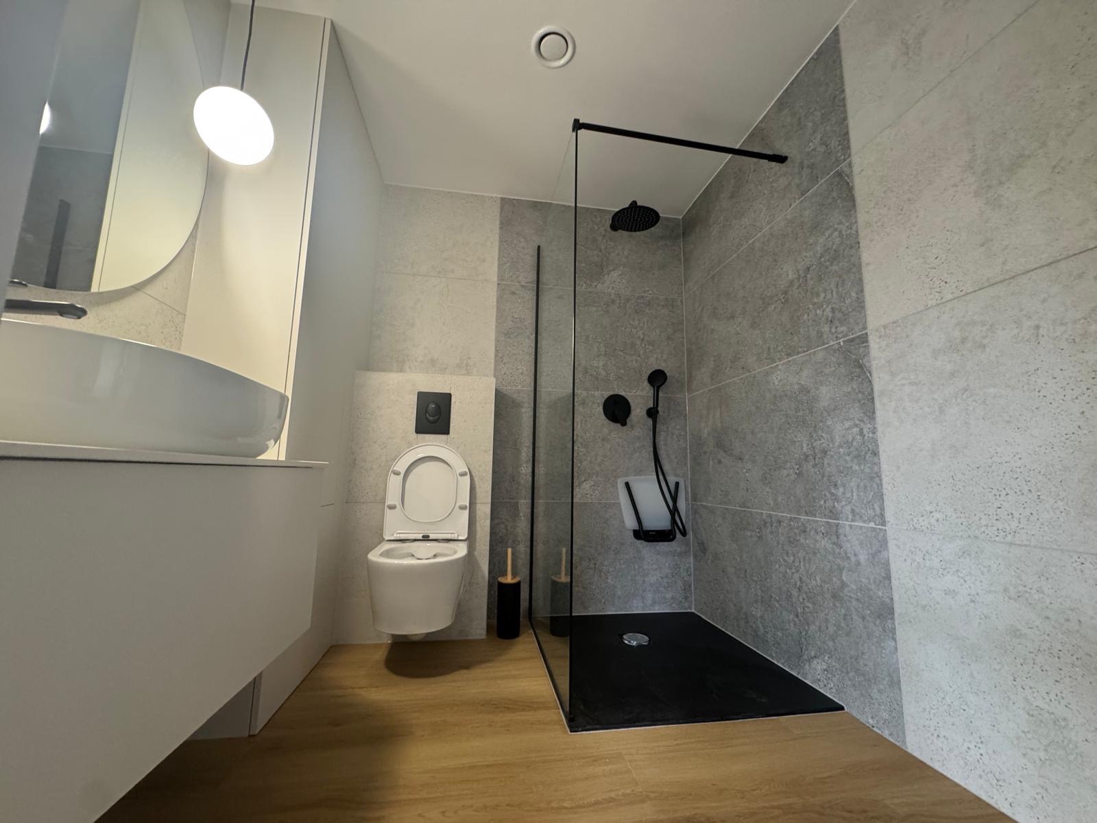 Nowoczesna łazienka z kabiną typu walk-in, szare płytki, drewnopodobna podłoga, biała umywalka i toaleta podwieszana. Minimalistyczny design z czarnymi akcentami.