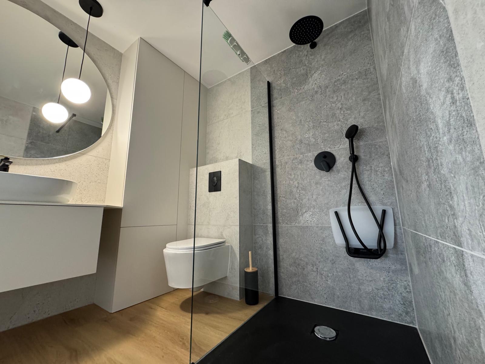 Nowoczesna łazienka z szarą glazurą, czarną armaturą i kabiną prysznicową typu walk-in. Umywalka z okrągłym lustrem i drewnopodobna podłoga dodają ciepła.