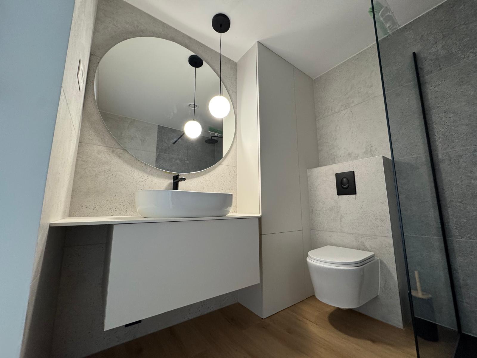 Nowoczesna łazienka z owalnym lustrem, białą umywalką nablatową i podwieszaną toaletą. Szare płytki imitujące beton, drewnopodobna podłoga i minimalistyczne oświetlenie.