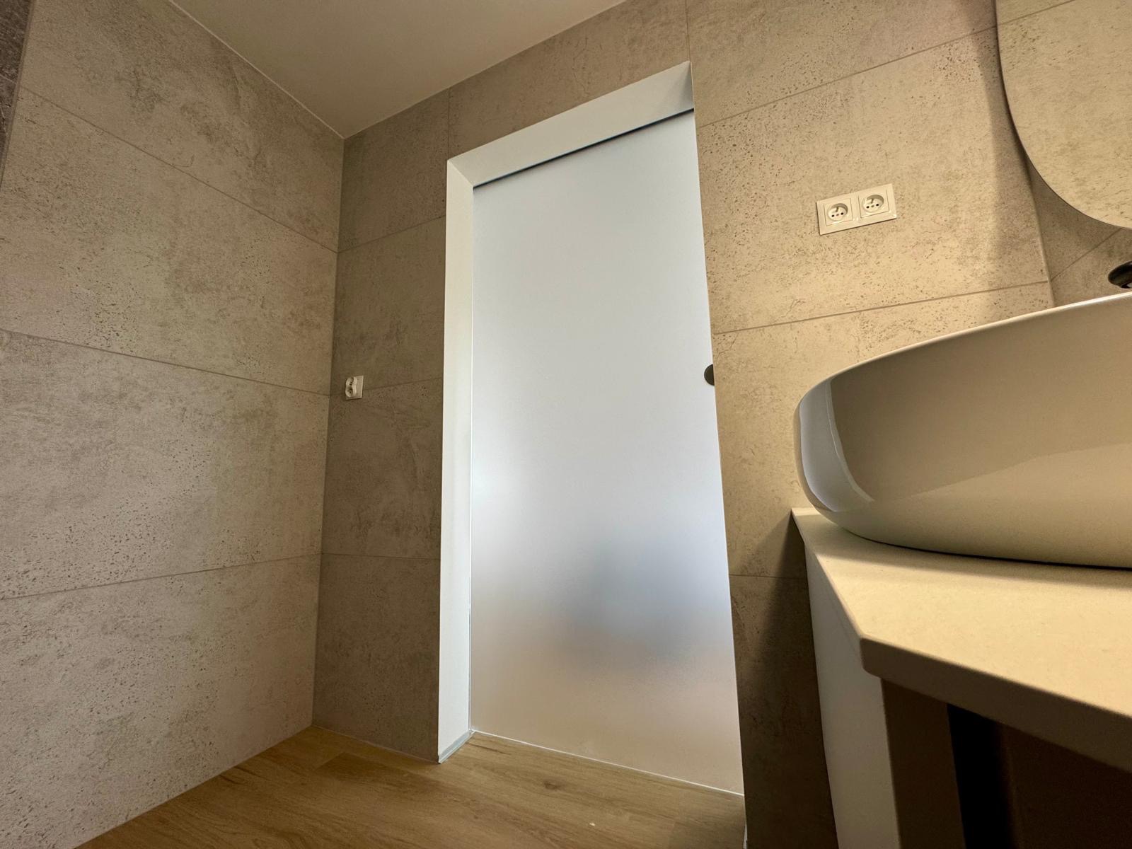 Nowoczesna łazienka z umywalką nablatową, płytkami imitującymi beton i przesuwnymi drzwiami z matowego szkła. Widoczne gniazdka elektryczne.