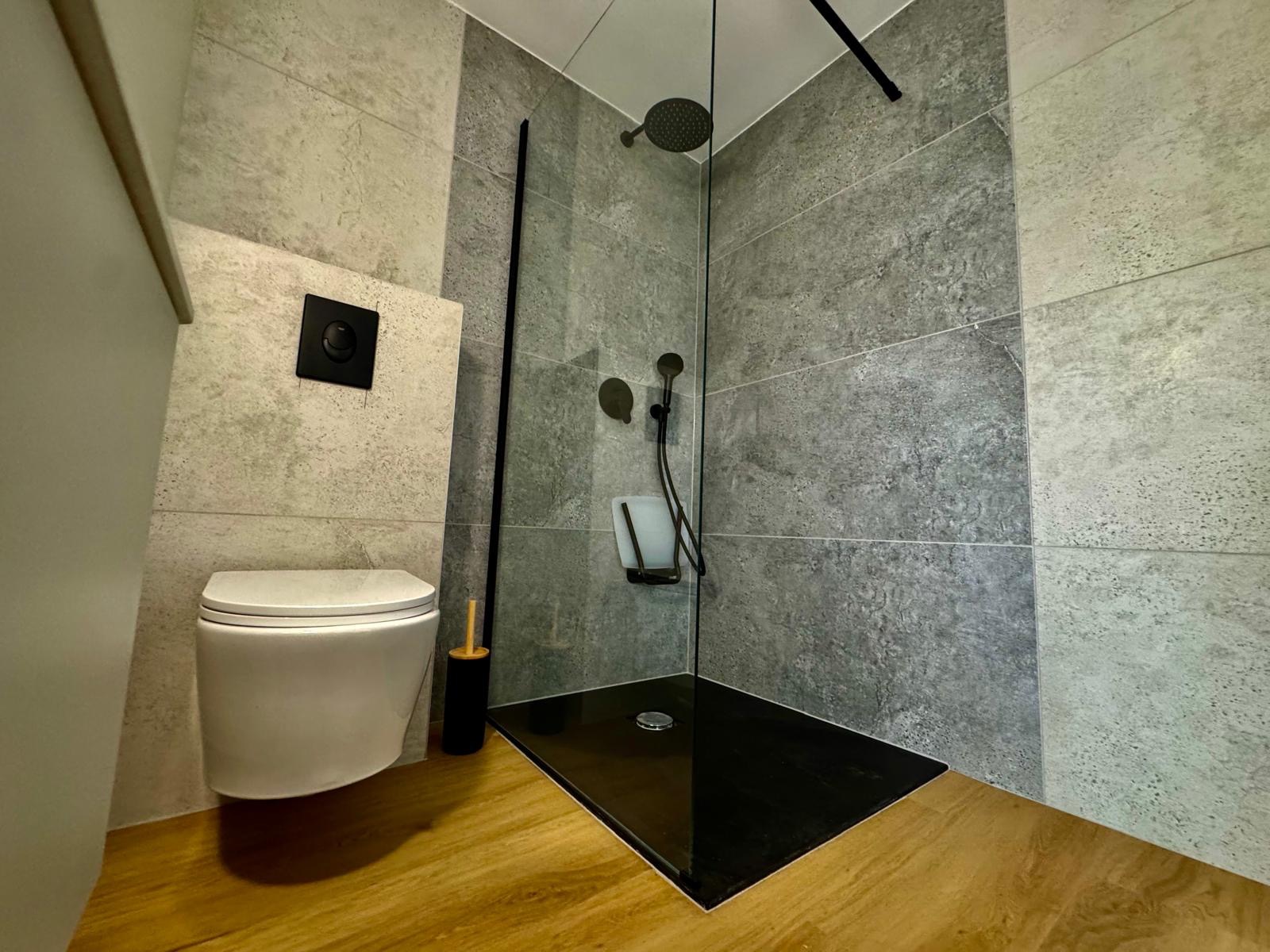 Nowoczesna łazienka z kabiną typu walk-in, szare płytki imitujące beton, podłoga drewnopodobna, wisząca toaleta i czarne akcenty.