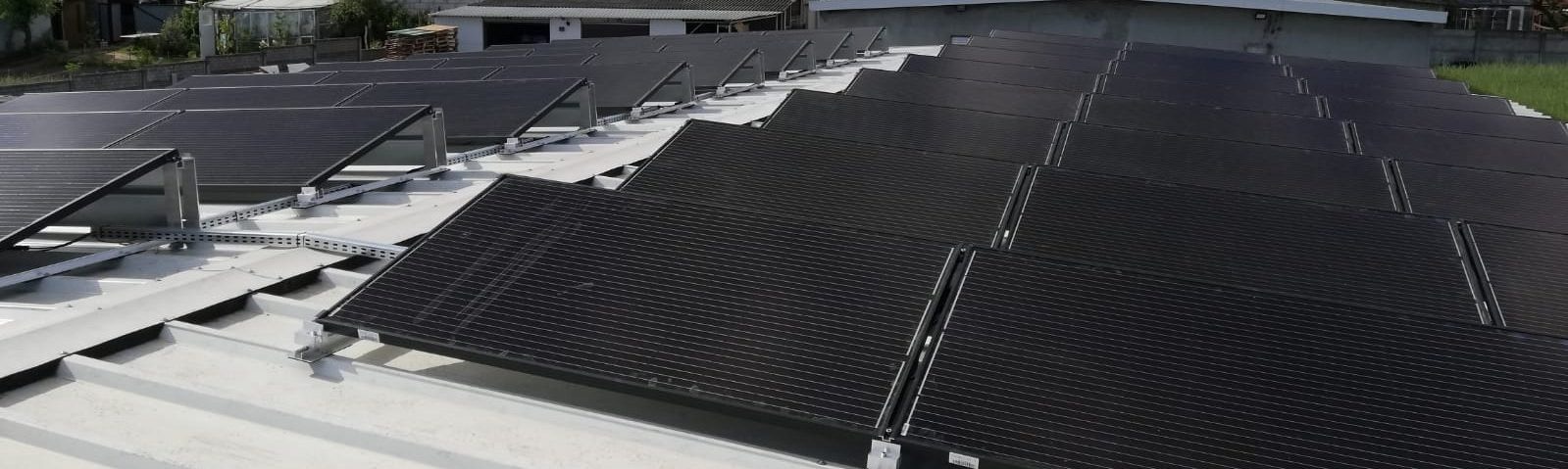 Rząd paneli fotowoltaicznych zamontowanych na dachu budynku, widoczne metalowe konstrukcje mocujące, w tle zielona roślinność i budynki gospodarcze.