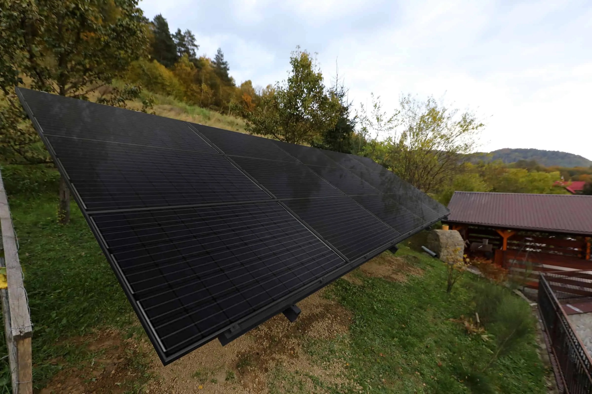 Czarny panel fotowoltaiczny na zboczu w Nowej Wsi, zainstalowany na metalowej konstrukcji, z widokiem na zielone wzgórza i budynek z brązowym dachem w tle.