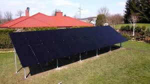 Instalacja fotowoltaiczna na metalowej konstrukcji, widoczna z góry na tle trawnika i domu z czerwonym dachem, z elementami krajobrazu w tle.