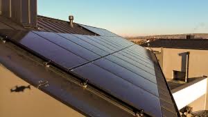 Panele fotowoltaiczne zamontowane na ciemnym, spadzistym dachu domu jednorodzinnego, widoczne z perspektywy drona rzucającego cień na elewację.