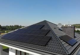 Panele fotowoltaiczne zamontowane na dachu domu jednorodzinnego z czarną dachówką, widoczne również komin i antena satelitarna na tle błękitnego nieba.