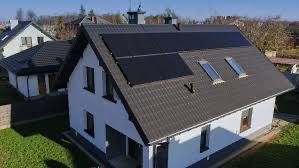 Biały dom jednorodzinny z ciemnoszarą dachówką i panelami fotowoltaicznymi na dachu, widok z góry w słoneczny dzień.