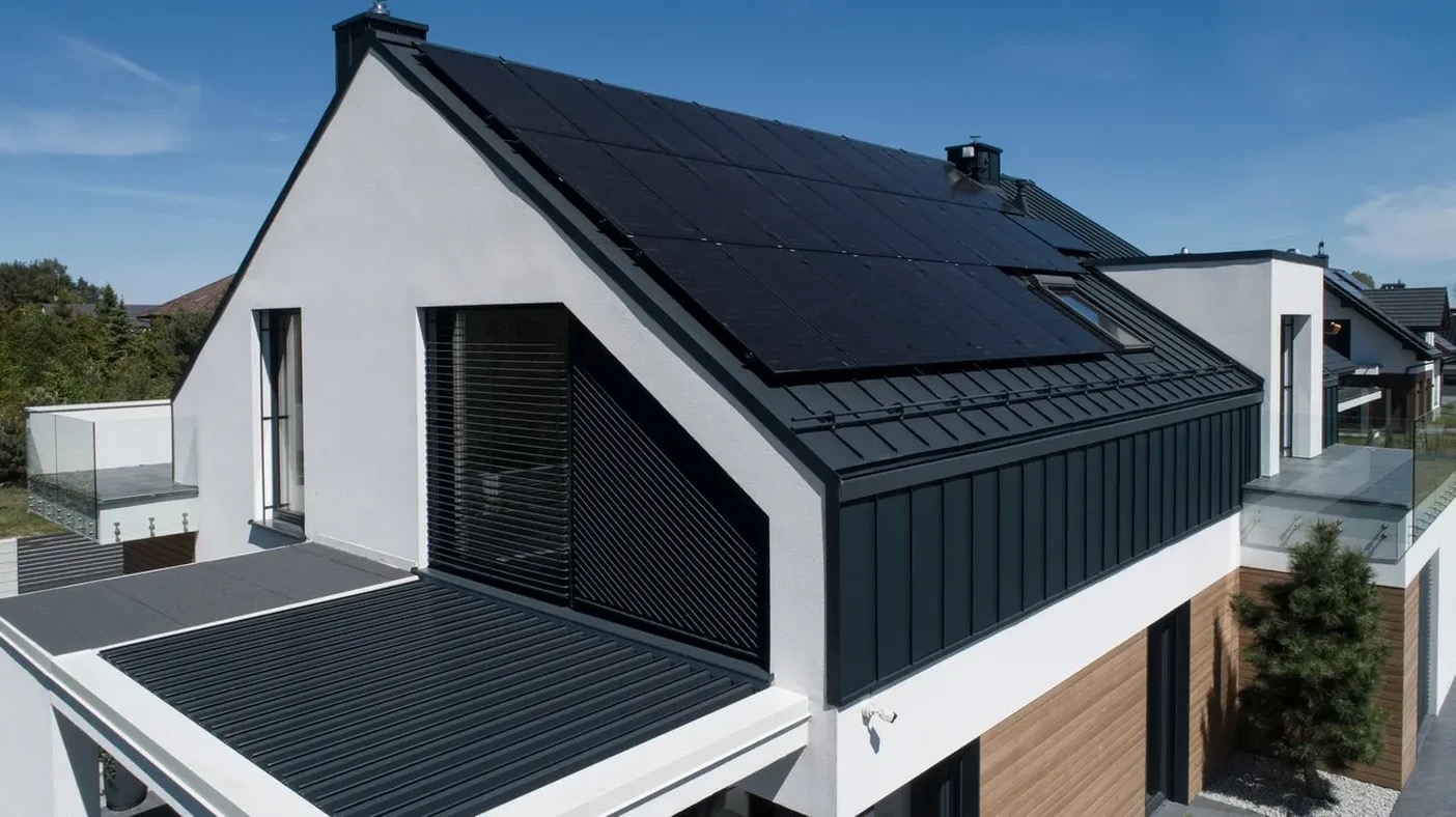 Nowoczesny dom jednorodzinny z panelami słonecznymi na dachu, widok z góry na białą elewację, szare elementy wykończeniowe i przeszklony balkon.