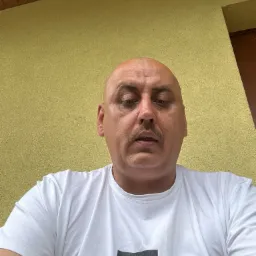 Portret mężczyzny w białej koszulce z logo Hugo Boss na tle żółtej, teksturowanej ściany. Ujęcie z dołu, widoczna część sufitu.