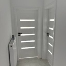 Piotr Łukaszewicz - Białe, nowoczesne drzwi z przeszkleniami w wąskim korytarzu po remoncie. Minimalistyczny design, świeżo pomalowane ściany i grzejnik.