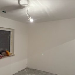 Piotr Łukaszewicz - Białe, świeżo pomalowane ściany w pokoju z oknem. Widoczne ślady po remoncie na podłodze i niedokończone wykończenie przy oknie. Oświetlenie sufitowe.