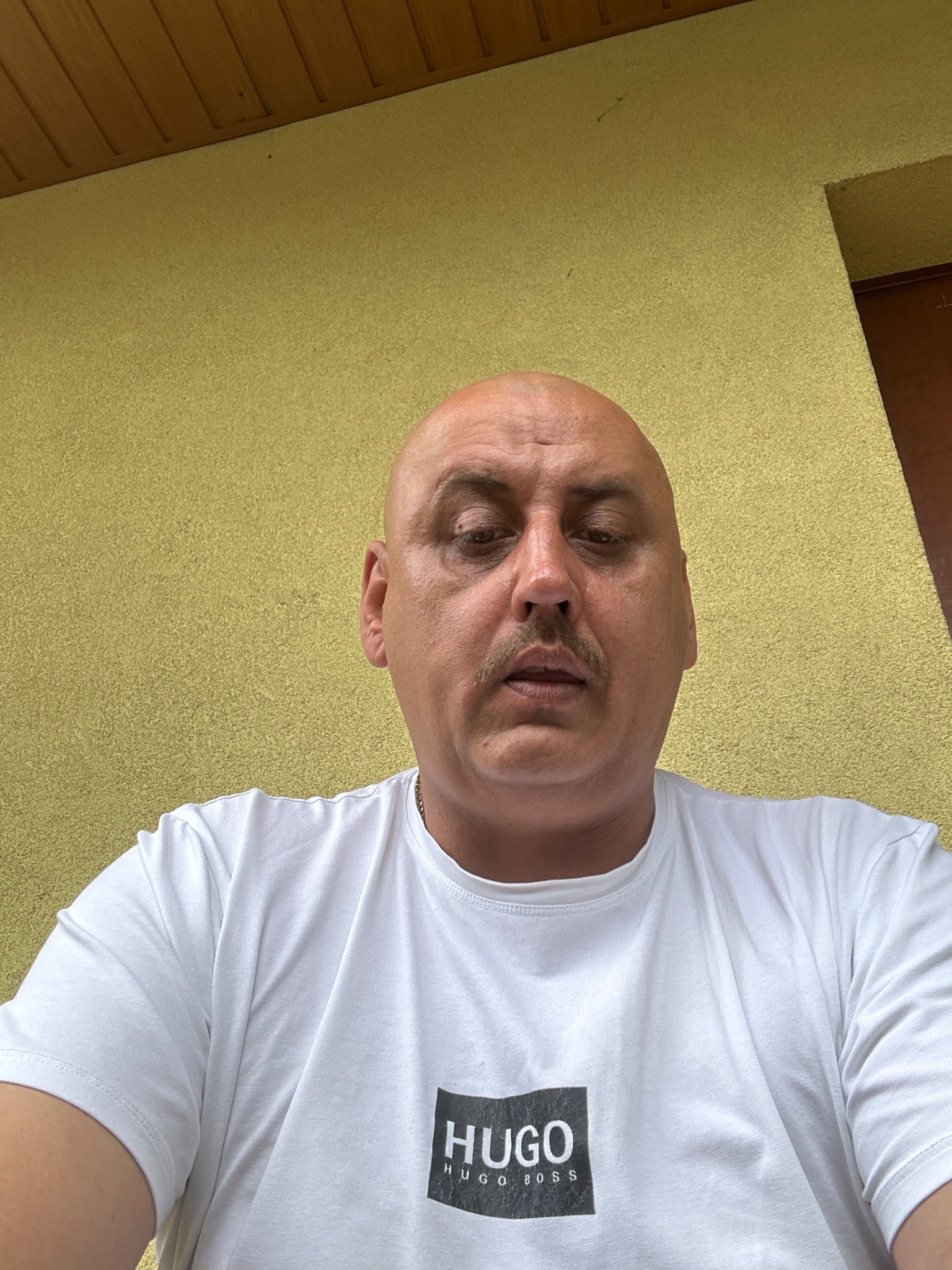 Portret mężczyzny w białej koszulce z logo Hugo Boss na tle żółtej, teksturowanej ściany. Ujęcie z dołu, widoczna część sufitu.