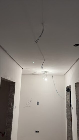 Białe wnętrze w trakcie remontu: gładzie na ścianach i suficie, widoczne kable elektryczne, surowy stan wykończenia otworów drzwiowych. Oświetlenie tymczasowe.