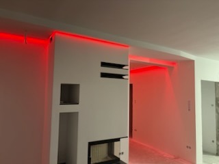 Biała ściana z wnękami i kominkiem, oświetlona czerwonym światłem LED. Widoczne przejście do innego pomieszczenia również z czerwonym oświetleniem.