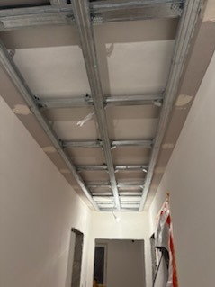 Sufit podwieszany w trakcie montażu: metalowa konstrukcja i płyty gipsowe, widok z dołu w korytarzu. Etap prac wykończeniowych.