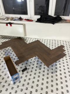 Układanie paneli w jodełkę na podłodze w pokoju. Widoczne narzędzia i karton po panelach. Białe okno w tle. Etap prac wykończeniowych.