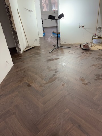 Ułożona, ciemna podłoga drewniana w jodełkę w trakcie prac wykończeniowych w Wołominie. Widoczne lampy budowlane i ślady po pracach remontowych.