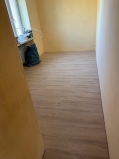 Jasne panele podłogowe w pokoju po remoncie, widok zza framugi. Ściany w odcieniach beżu i żółci, okno z workiem na śmieci w tle. Nowa podłoga po montażu.