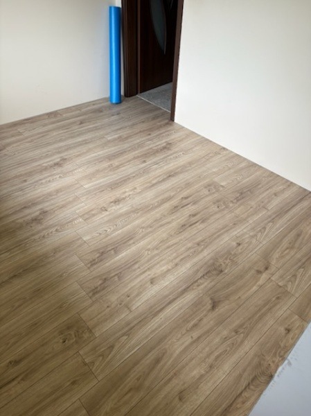 Ułożone panele podłogowe w jasnym odcieniu drewna, widoczne przy ścianie i progu drzwi. W rogu niebieska rolka. Minimalistyczne wnętrze po remoncie.