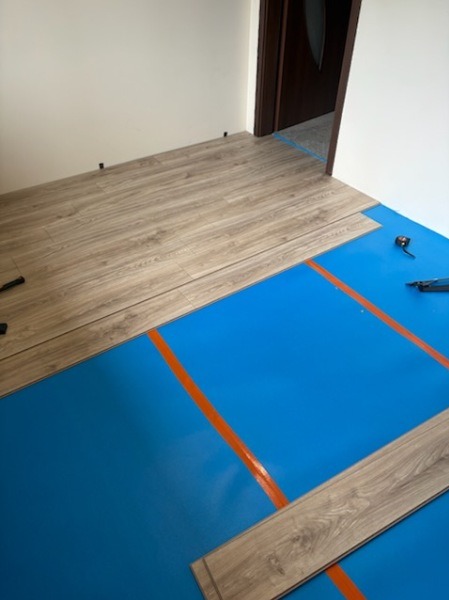 Układanie paneli podłogowych na niebieskiej folii izolacyjnej. Widoczne narzędzia i fragment ułożonej podłogi przy ścianie. Jasne panele imitujące drewno.