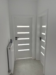 Białe, nowoczesne drzwi z przeszkleniami w wąskim korytarzu po remoncie. Minimalistyczny design, świeżo pomalowane ściany i grzejnik.