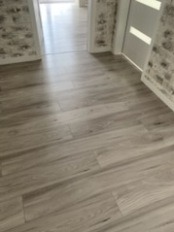 Ułożone panele podłogowe w odcieniach szarości, imitujące drewno, w korytarzu z tapetą w cegiełki i białymi drzwiami. Widok z perspektywy podłogi.
