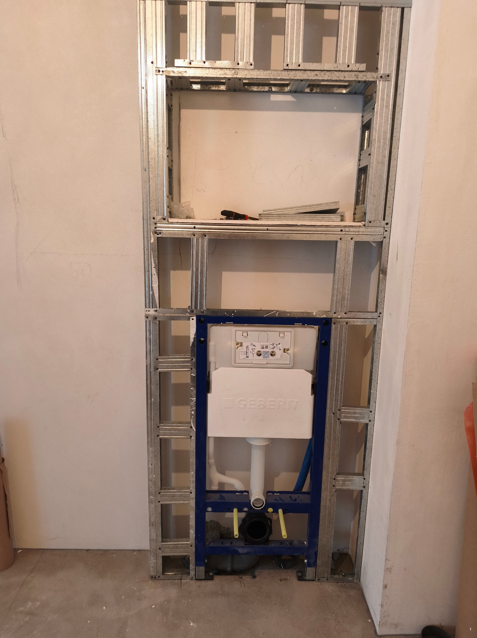 Stelaż podtynkowy Geberit zamontowany w metalowej konstrukcji przy ścianie. Widoczne elementy instalacji wodno-kanalizacyjnej. Etap prac wykończeniowych w łazience.