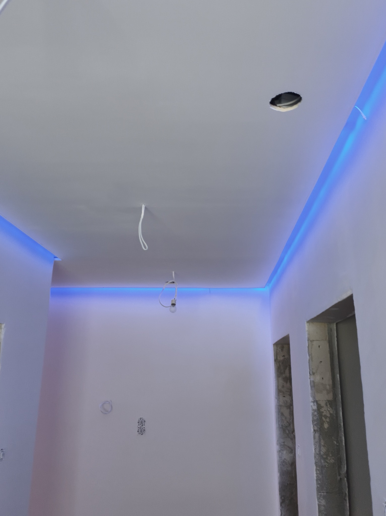 Białe wnętrze z niebieskim oświetleniem LED na suficie, widoczne kable i otwór na lampę, surowe wykończenie ościeżnic drzwiowych.